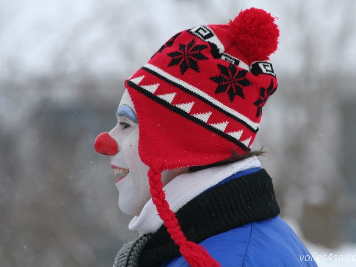 Clown d’hiver vu de profil, portant une tuque rouge et un costume chaud, dans un décor extérieur hivernal