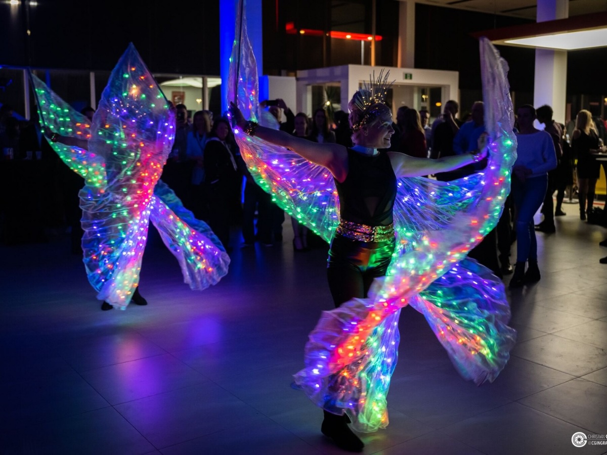Deux artistes en costumes lumineux élégants dotés d’ailes articulées en mouvement, évoluant en animation ambulatoire devant une foule dans l’ombre