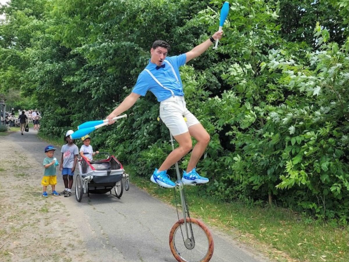 Amuseur en équilibre sur un monocycle girafe, tenant trois quilles de jonglerie bleues dans ses mains