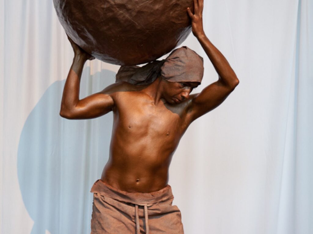 Statue vivante entièrement peinturée en bronze, portant une grosse sphère sur l’épaule dans une posture rappelant Atlas