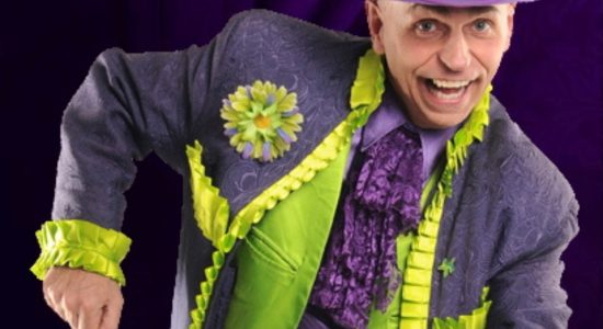 Personnage souriant vêtu d’un costume mauve et vert fluo, en pleine animation dansante